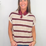 A smiling woman wearing a short-sleeve knit top with horizontal burgundy and cream stripes, paired with a burgundy neck scarf and blue jeans, standing against a plain white wall.