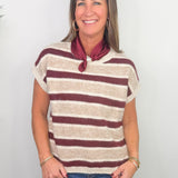 A smiling woman wearing a short-sleeve knit top with horizontal burgundy and cream stripes, paired with a burgundy neck scarf and blue jeans, standing against a plain white wall.