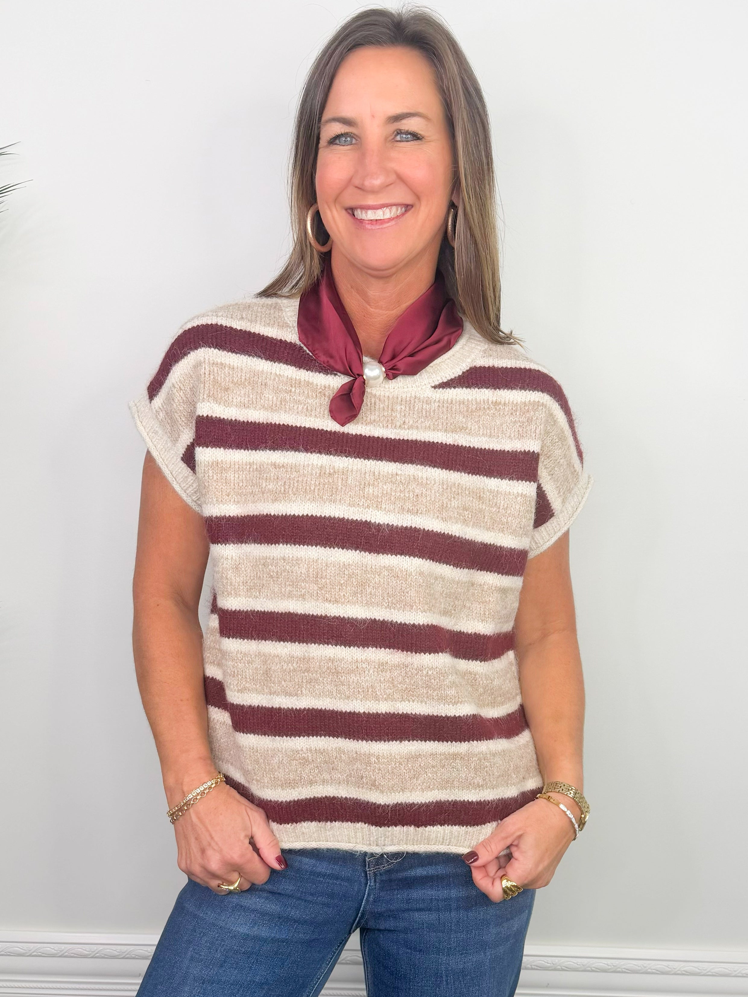 A smiling woman wearing a short-sleeve knit top with horizontal burgundy and cream stripes, paired with a burgundy neck scarf and blue jeans, standing against a plain white wall.
