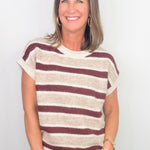 A smiling woman with shoulder-length brown hair wearing a short-sleeve knit top with horizontal burgundy and cream stripes, paired with blue jeans, standing against a plain white background.