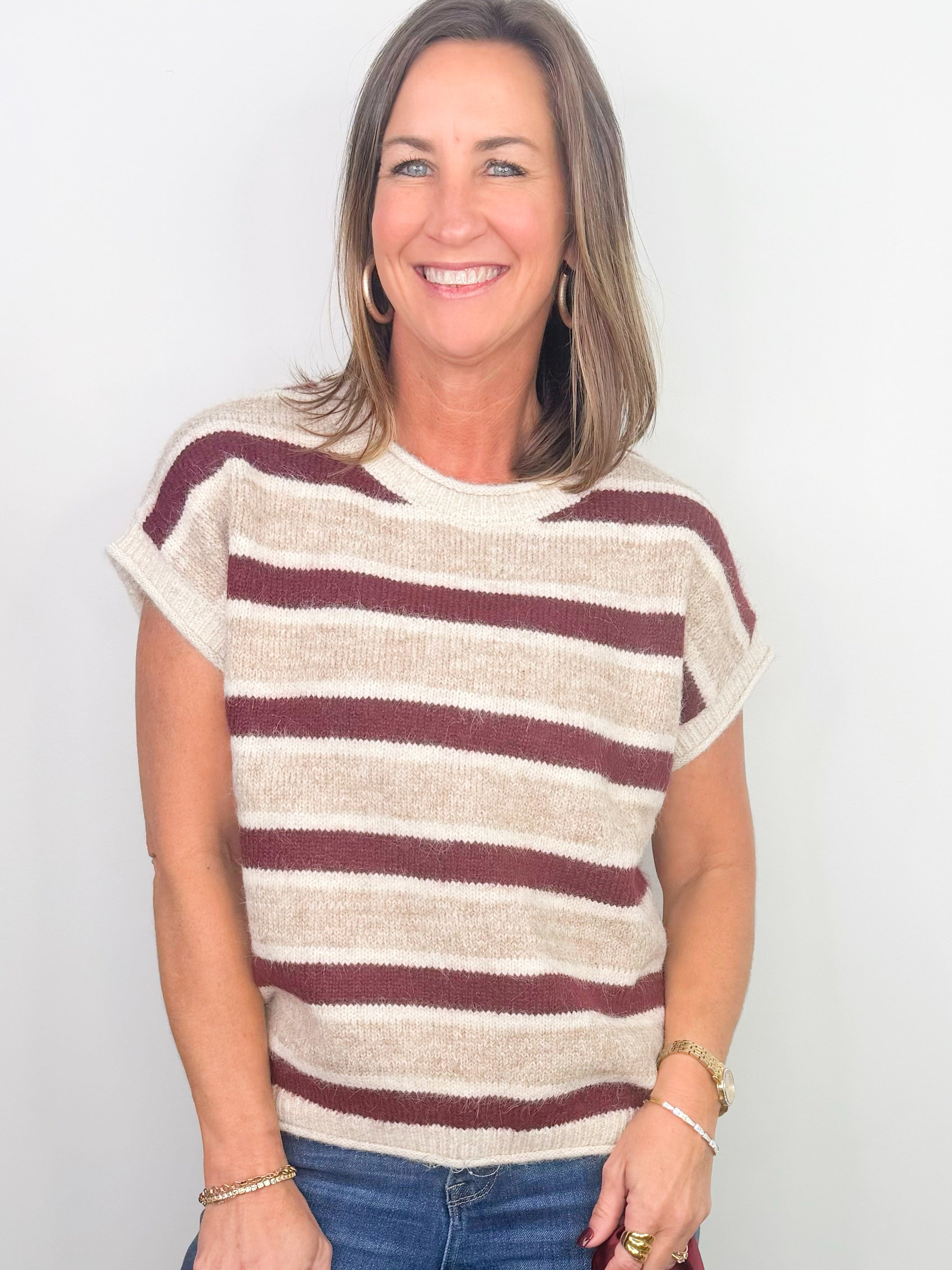 A smiling woman with shoulder-length brown hair wearing a short-sleeve knit top with horizontal burgundy and cream stripes, paired with blue jeans, standing against a plain white background.