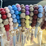A close-up of a person's hands holding a stack of colorful beaded bracelets and pen-style keychains inside a car. The beads come in multiple pastel and bright colors including blue, pink, red, purple, yellow, brown, and white, with some featuring natural wood geometric accents and metallic keychains.